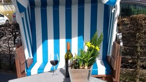A blue and white striped awning, a table with a wine bottle and a potted plant, and a parked car in the background. - Haffblick Whg. Ha18, Deutschland (Ostseebad Boltenhagen)