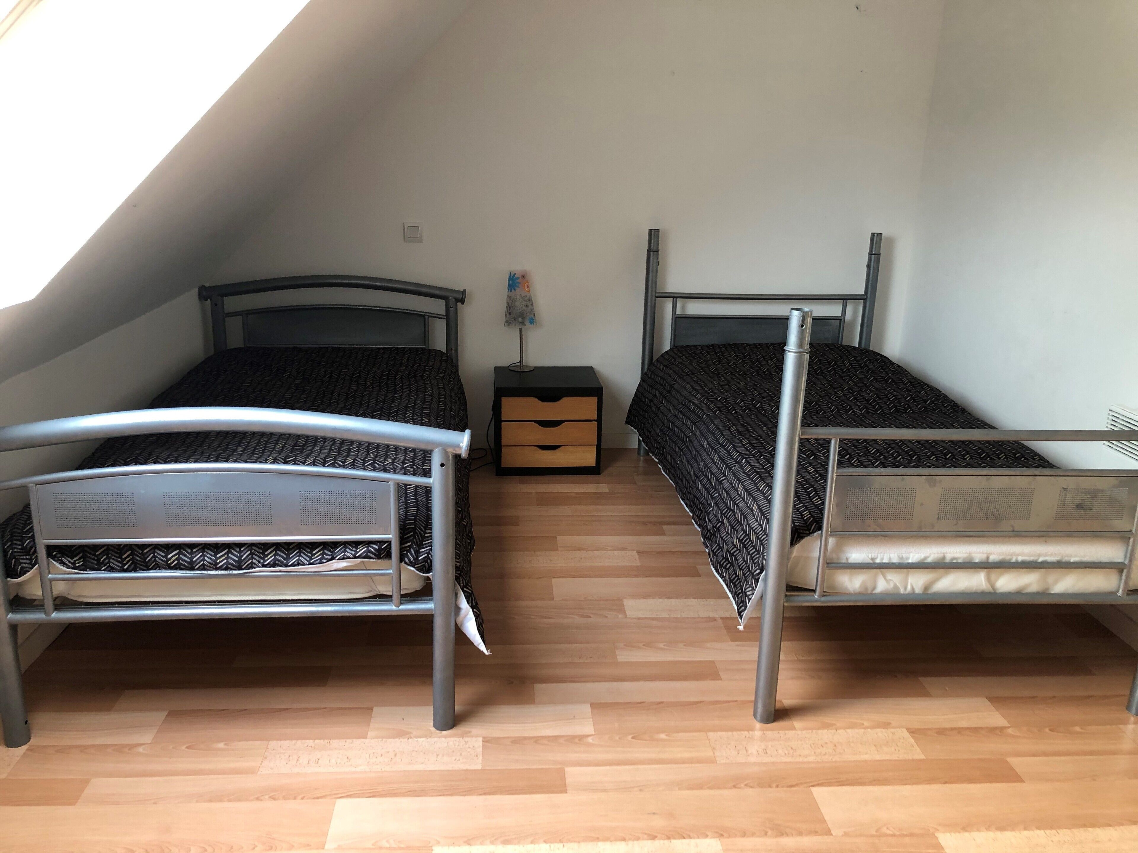 Une chambre de dortoir avec deux lits superposés en métal, une table de chevet avec des tiroirs, une lampe et un plafond en pente.