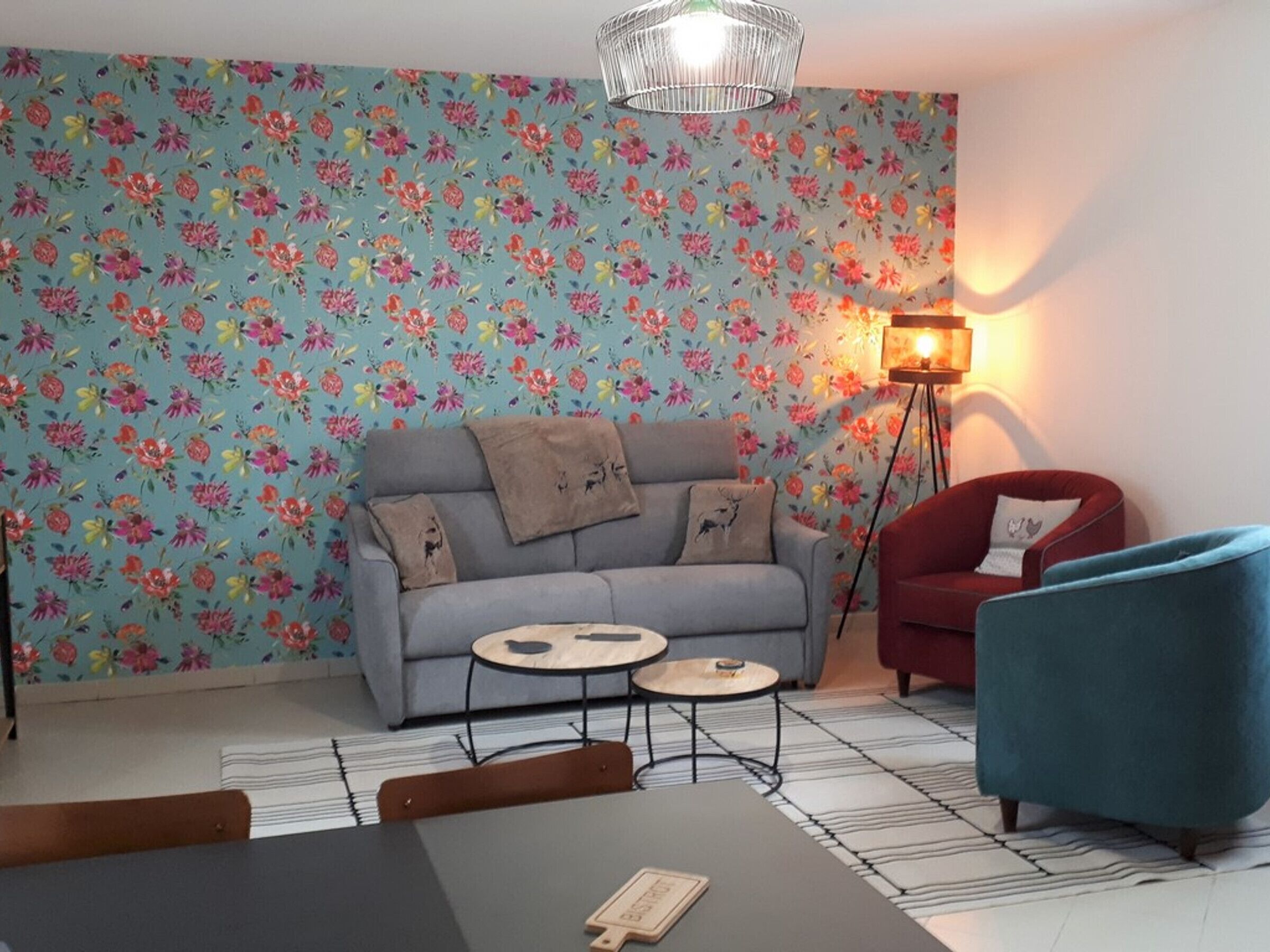 Una sala de estar acogedora con papel tapiz floral, un sofá gris, un sillón rojo, un sillón azul, una mesita de centro redonda y una lámpara de pie.