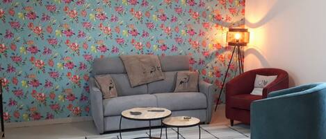 Una sala de estar acogedora con papel tapiz floral, un sofá gris, un sillón rojo, un sillón azul, una mesita de centro redonda y una lámpara de pie.