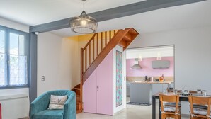 A modern living room with a blue sofa, a wooden dining table with chairs, a kitchen area with pink cabinets, and a staircase with wooden railings. - Charming Getaway with Garden, WiFi, and Garage in Scenic Gouy-les-Groseillers (Gouy-les-Groseillers)