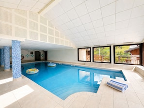 Ein Hallenbad mit klarem, blauem Wasser, Fliesenboden und Blick auf eine grüne Landschaft durch große Fenster.