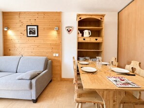 Ein gemütliches Zimmer mit Holzwand, einer grauen Couch, einem Holztisch gedeckt mit Tellern, Gläsern und Besteck sowie einem Regal mit Küchenutensilien.
