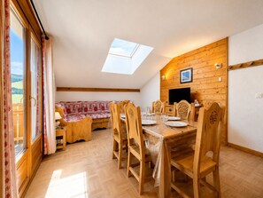A dining area with a wooden table and chairs, a sofa, and a television on the wall.