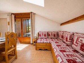A room with a wooden floor, a sofa with patterned cushions, a wooden chair, a table with chairs, and a door leading outside.