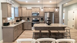 A modern kitchen with wooden cabinets, a central island with bar stools, a microwave, a refrigerator, and pendant lighting. - Luxury 7BR Reunion Lodge on Thousand Hills Golf Course | Sleeps 22 | Near Highway 76 (Branson)