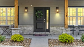 Una casa con puerta frontal oscura, dos columnas de madera, base de piedra, una corona decorativa y dos luces fijadas a la pared.