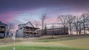 Campo de golf con un asta de bandera en primer plano, rodeado de edificios residenciales con balcones y un cielo despejado.