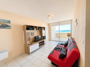 Ein Zimmer mit einem roten Sofa, einem Fernseher auf einem Holzständer, einem Balkon mit Meerblick und einem Fliesenboden.