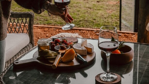 Una mesa dispuesta con queso, frutas y copas de vino, mientras una persona sirve vino.
