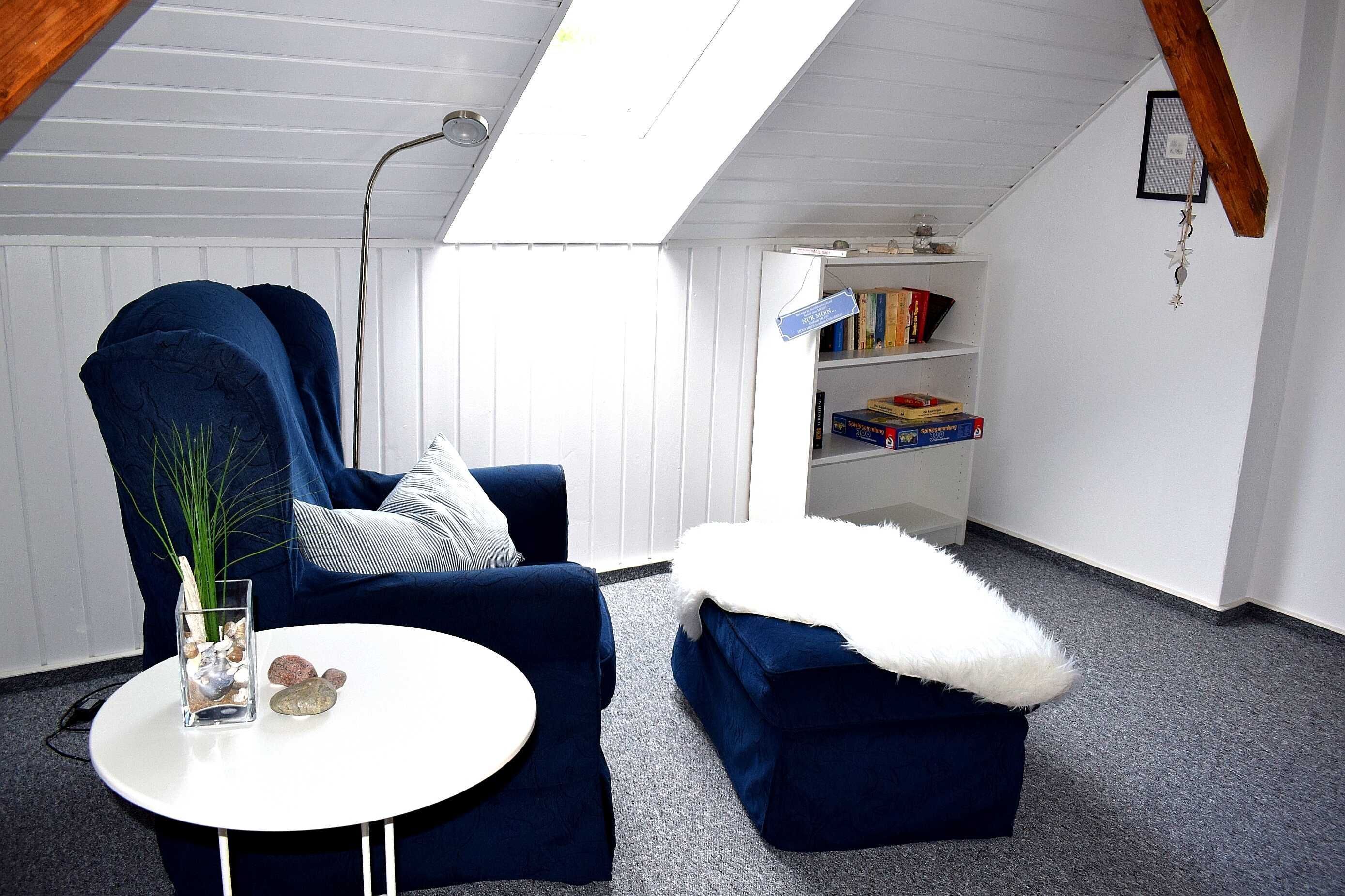 A room with a blue armchair, a white round table, a white fluffy ottoman, a bookshelf, and a skylight.