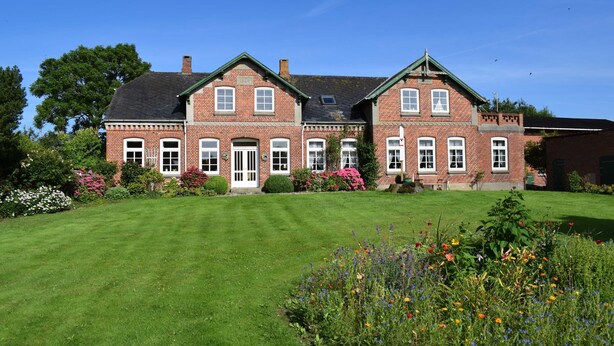 A large brick building with a symmetrical facade, multiple windows, and a well-maintained lawn with colorful flowers. - Ferienwohnung Andresen - FW Andresen (Pommerby)