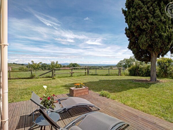 A deck with outdoor seating, a wooden fence, and a view of a grassy field and distant hills.