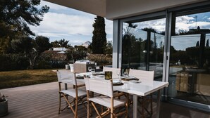 Un área de comedor exterior moderna con una mesa y sillas de vidrio, ubicada en una terraza de madera con vistas al paisaje circundante.