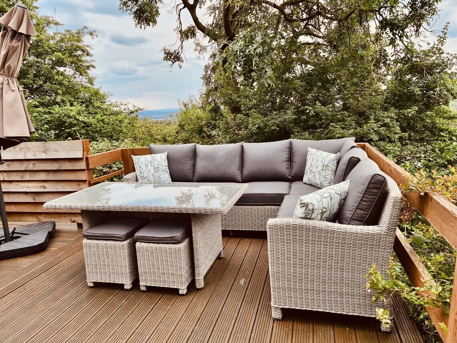 Un espace de détente extérieur aménagé avec des meubles en osier, une table en verre et des coussins, entouré d’une terrasse en bois et d’une végétation luxuriante.