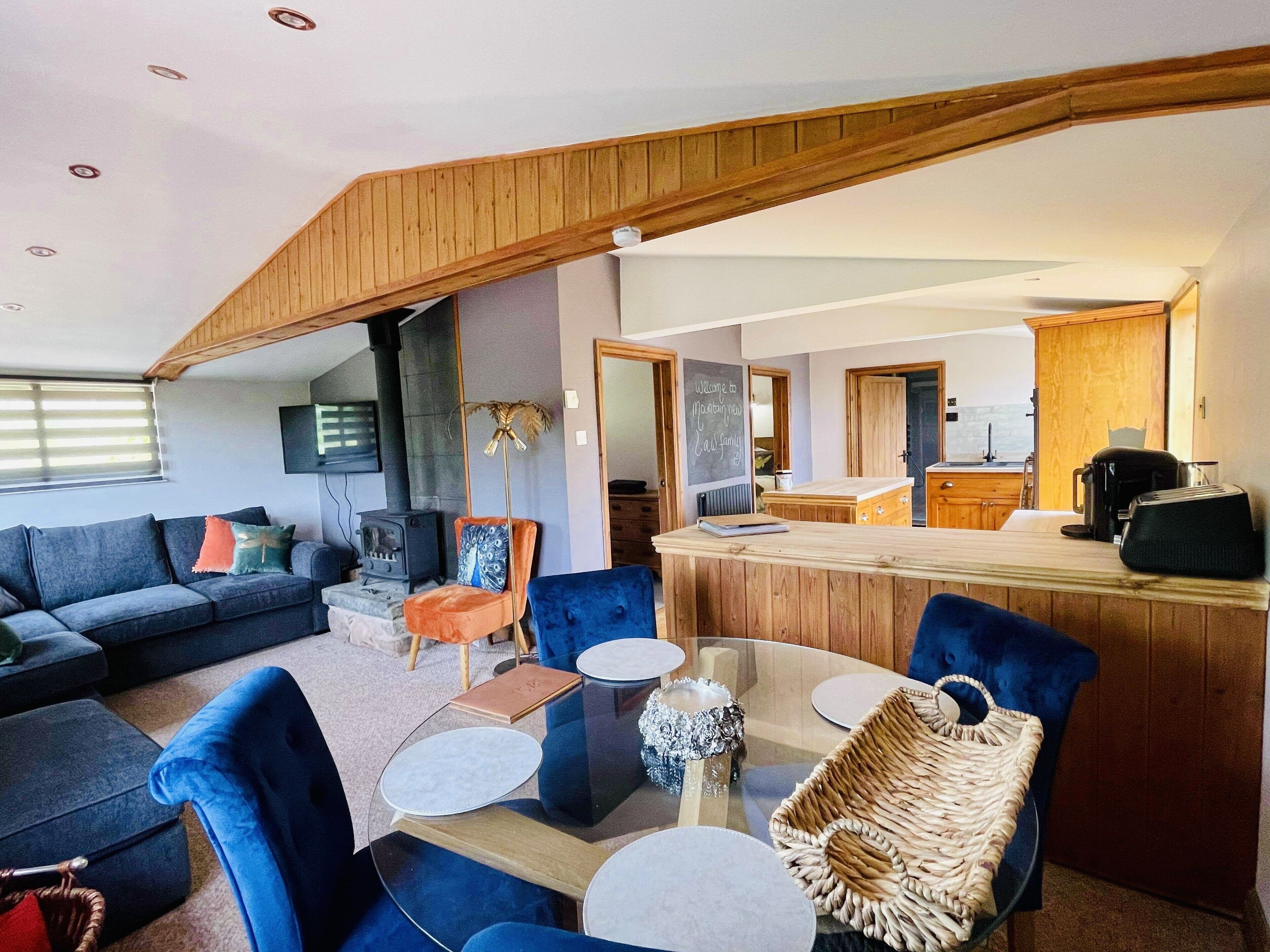 Un salon moderne avec un plafond haut, une cheminée, un coin repas avec une table en verre et une cuisine à l’arrière-plan.