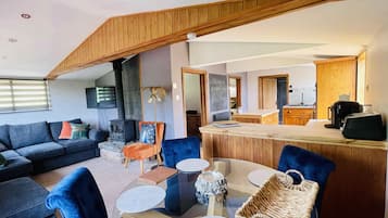 Un salon moderne avec un plafond haut, une cheminée, un coin repas avec une table en verre et une cuisine à l’arrière-plan.