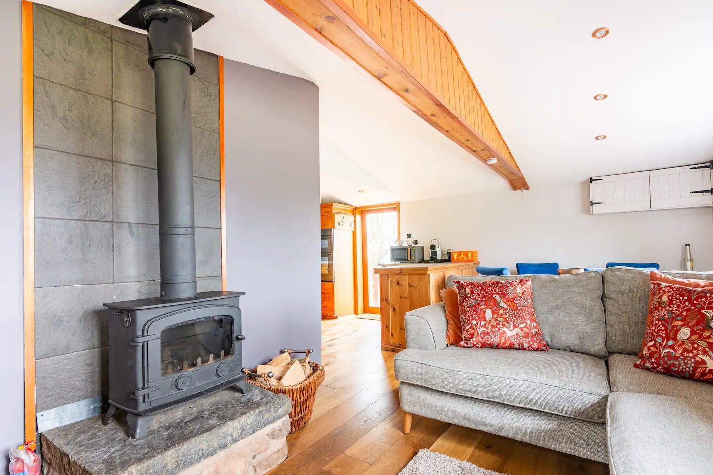 A cozy living room with a stone fireplace, a grey sofa, and a kitchen in the background.