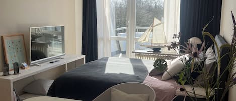 A modern hotel room with a bed, bedside table, TV, and a large window with curtains.