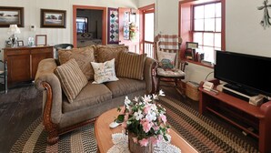 Un salon chaleureux avec un canapé marron, une table basse en bois ornée d’un vase de fleurs, un téléviseur posé sur un meuble et une fenêtre à cadre rouge.