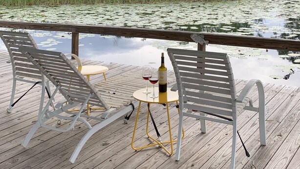 Una terraza de madera con dos sillas blancas y una pequeña mesa redonda, una botella de vino y copas sobre la mesa.