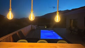 Een poolbar met hangende lampen, stoelen en uitzicht op een zwembad en bergen in de schemering.