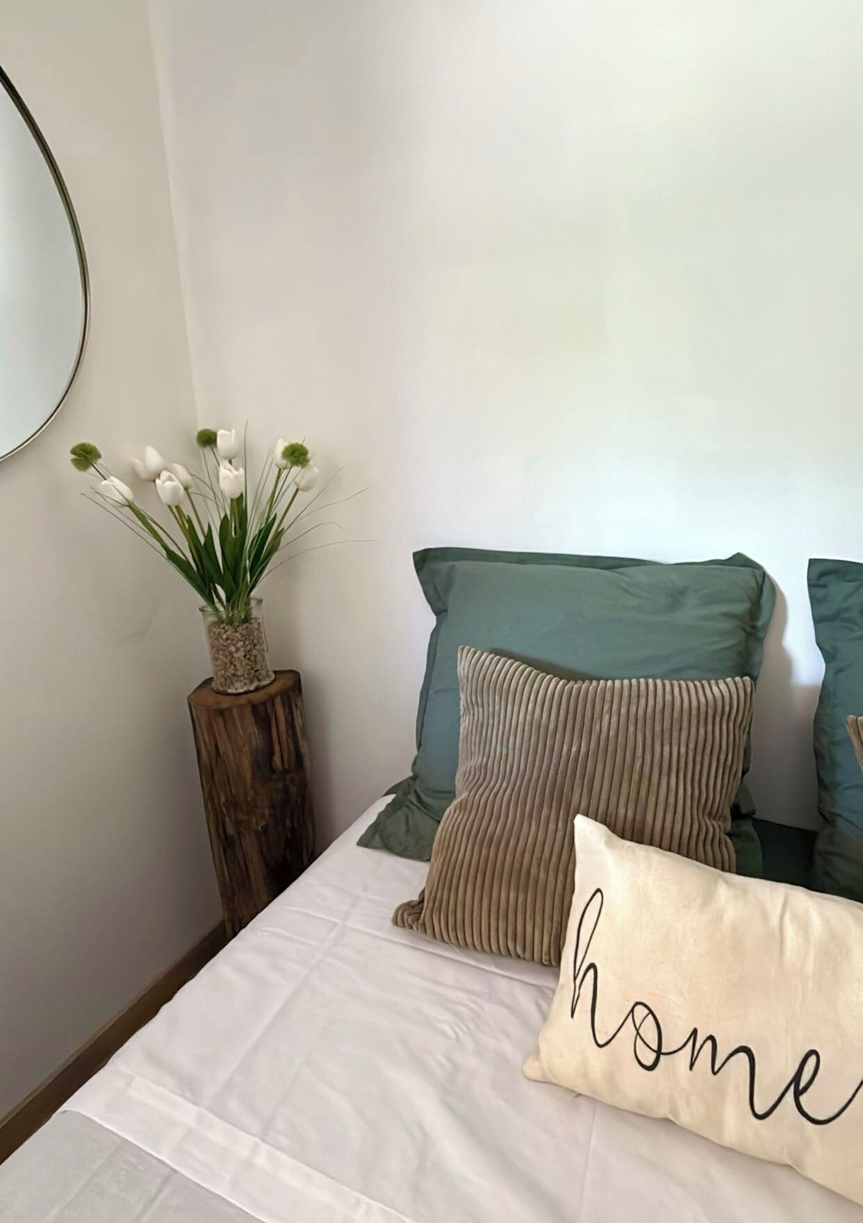 Ein Schlafzimmer mit einem Bett, Kissen, einer Vase mit Blumen und einem Spiegel an der Wand.