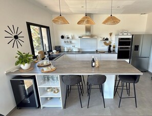 A modern kitchen with a central island, bar stools, pendant lights, and a built-in wine cooler.