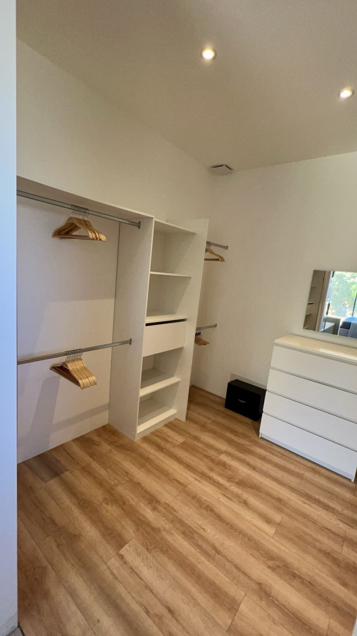 A modern, minimalist walk-in closet with open shelving and hanging space, wooden flooring, and a large mirror.