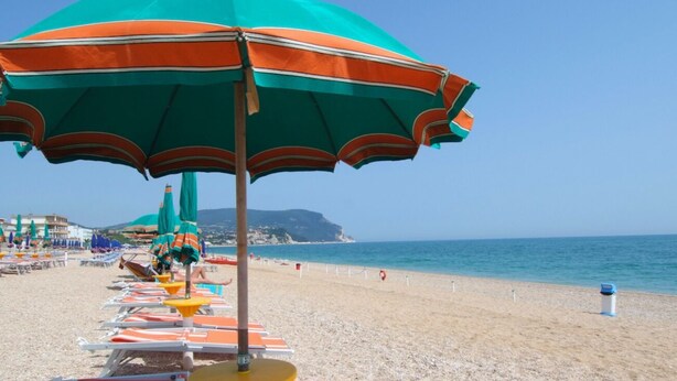Ein Strand mit Reihen von Liegestühlen und Sonnenschirmen, einem klaren Himmel und einer Küstenlinie im Hintergrund.