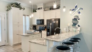 A modern kitchen with white cabinets, a black refrigerator, a microwave, and a coffee maker. There are three bar stools and a decorative wall piece. - Dauphin Tale - Family Friendly Gulf View Home only Steps to Gulf of Mexico (Dauphin Island)