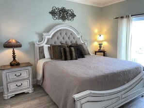 Un dormitorio con cabecera capitonada, cama con ropa de cama estampada, dos mesitas de noche con lámparas y un adorno en la pared.
