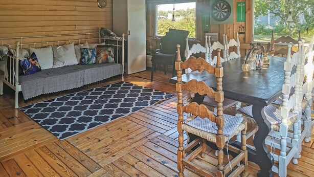 Interior acogedor de una cabaña con suelo de madera, un sofá con cojines estampados, una mesa de comedor con sillas y vistas a la vegetación exterior.