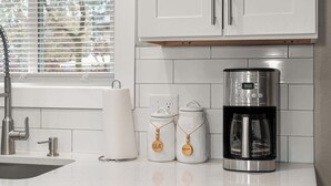 Una cocina con cafetera, dos frascos etiquetados 'SUGAR' y 'CREAM', y un dispensador de servilletas de papel.