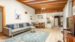 Una sala de estar con un sofá, una mesa de centro y una alfombra. Hay un comedor con una mesa y sillas. La habitación tiene vigas de madera en el techo y una zona de cocina con un refrigerador y armarios.