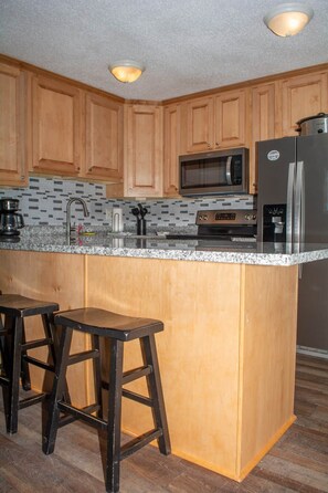 A kitchen with wooden cabinets, a granite countertop, a microwave, a refrigerator, and two bar stools. - Winterplace E103 Ski in Ski out condo First floor (Ghent)