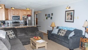 Ein Wohnzimmer mit grauem Sofa, einem Couchtisch und einer Küchenzeile mit Holzschränken und Geräten.