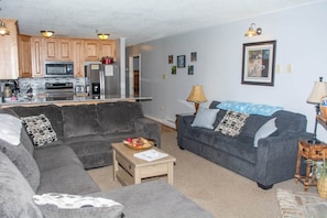 A living room with a grey sofa, a coffee table, and a kitchen area with wooden cabinets and appliances. - Winterplace E104 Ski in Ski out condo First floor (Ghent)