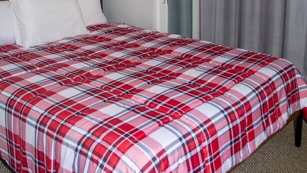 Ein Schlafzimmer mit einem Bett, weißen Kissen, einer rot-weißen Karodecke, einer Wandleuchte und einer Tür mit grauem Vorhang.