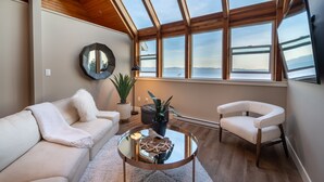 A modern living room with a glass coffee table, white sofa, and armchair, large windows with a view of the sea, and a wooden ceiling. - The Owl’s Nest | Panoramic Ocean View Retreat (Sooke)