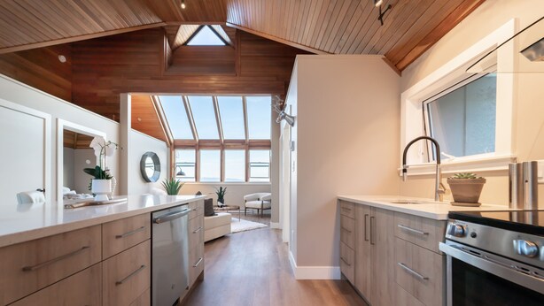 A modern kitchen with wooden ceiling beams, a large window, and a built-in oven. - The Owl’s Nest | Panoramic Ocean View Retreat (Sooke)