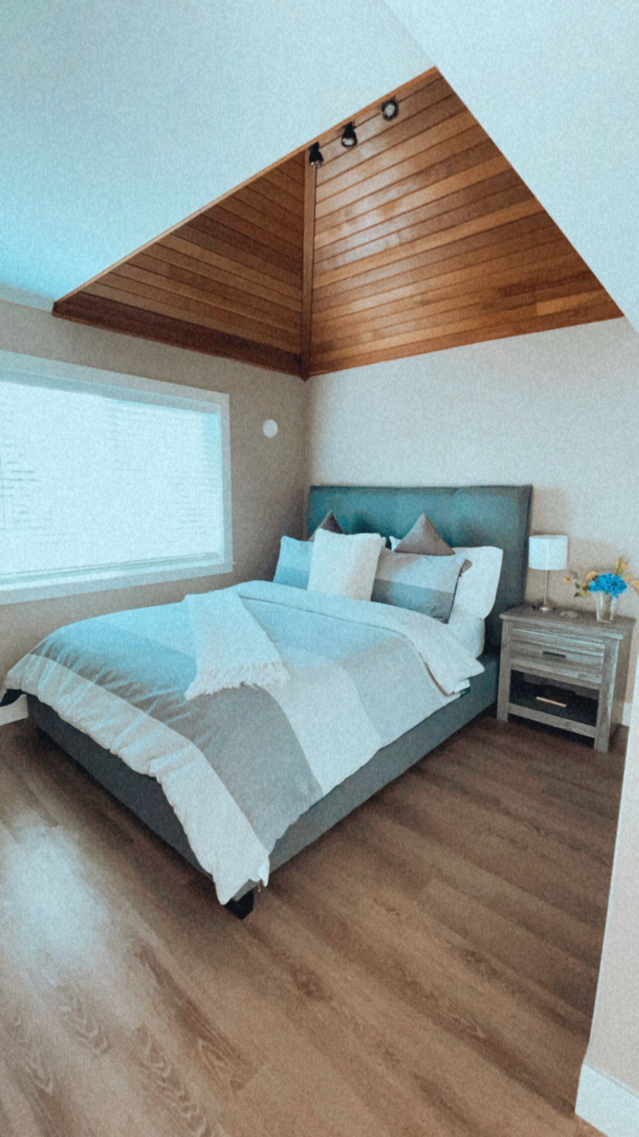 A bedroom with a sloped ceiling, a bed with a blue and white bedspread, a nightstand with a lamp, and a window with blinds.