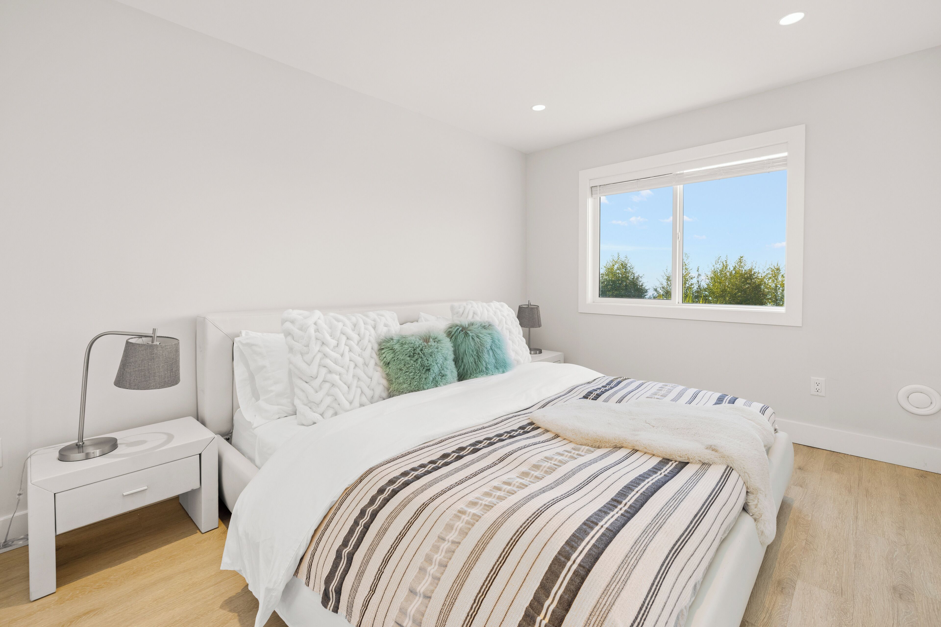 Quarto moderno com uma cama grande, mesa de cabeceira, abajur e janela com vista para o verde.