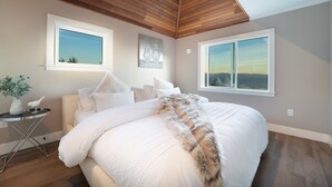 A bedroom with a large bed, two windows, a wooden ceiling, and a small side table with a plant. - The Owl’s Nest | Panoramic Ocean View Retreat (Sooke)