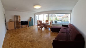 A spacious living room with a wooden parquet floor, a large window offering a view of buildings, a television, and a dining area with chairs and a table. - Studio Apartment Terrassengarten - Bad Kissingen (Bad Kissingen)