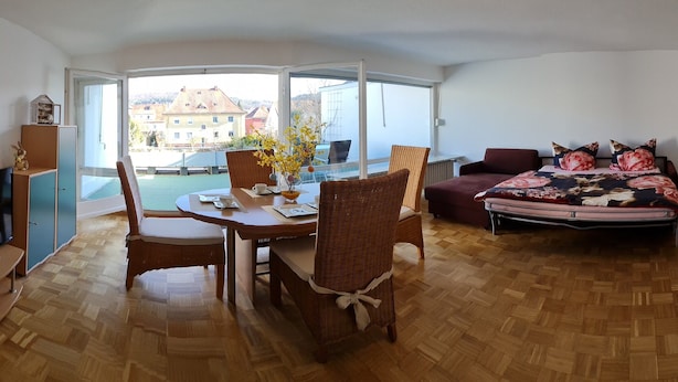 A room with a dining table, chairs, a television, a sofa, and a view of buildings through large windows. - Studio Apartment Terrassengarten - Bad Kissingen (Bad Kissingen)