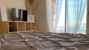 Une chambre à coucher avec un grand lit, un téléviseur posé sur une étagère en bois et une fenêtre ornée de rideaux légers.