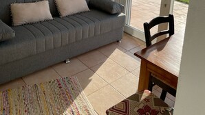 Un salón con un sofá gris, dos almohadas, una silla de madera, una mesita y vistas a una piscina a través de un ventanal.