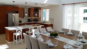 Una cocina moderna con una isla central, taburetes blancos tipo bar y una zona de comedor preparada para seis personas, con vajilla blanca.