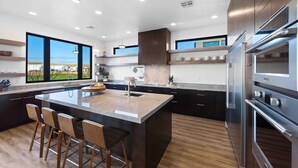 A modern kitchen with a large island, bar stools, stainless steel appliances, and large windows offering a view of the outdoors.